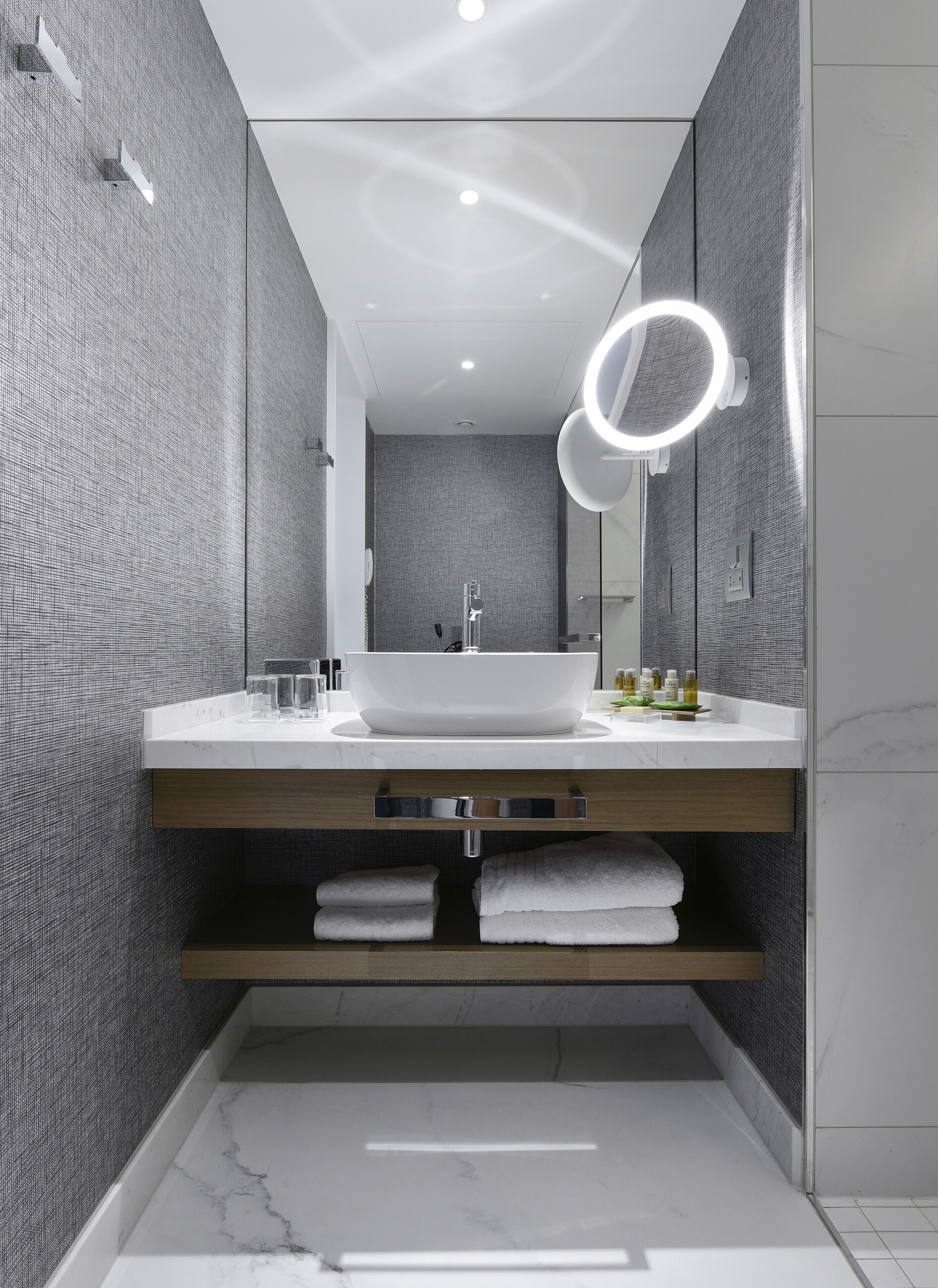 Un baño moderno con un lavamanos blanco grande, un espejo con luz circular y un perchero para toallas.