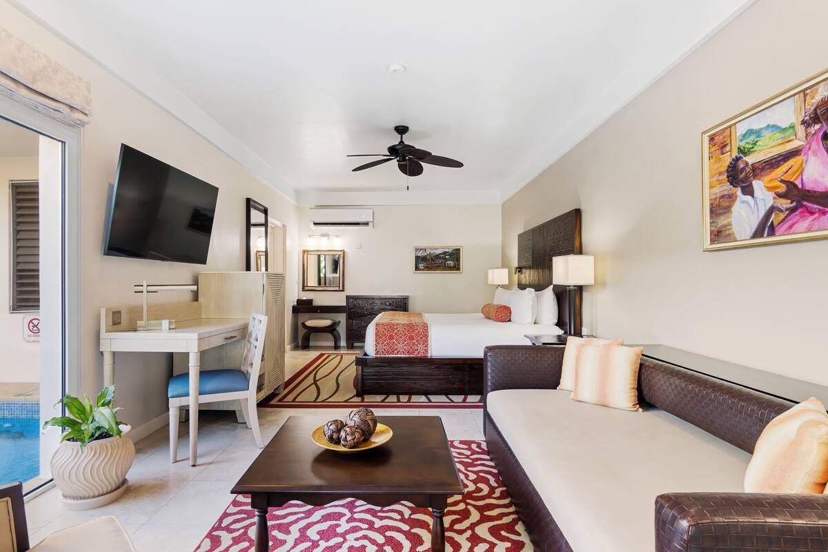 A hotel room with a bed, a sofa, a desk, a chair, a TV, a ceiling fan, a painting, and a view of a swimming pool.