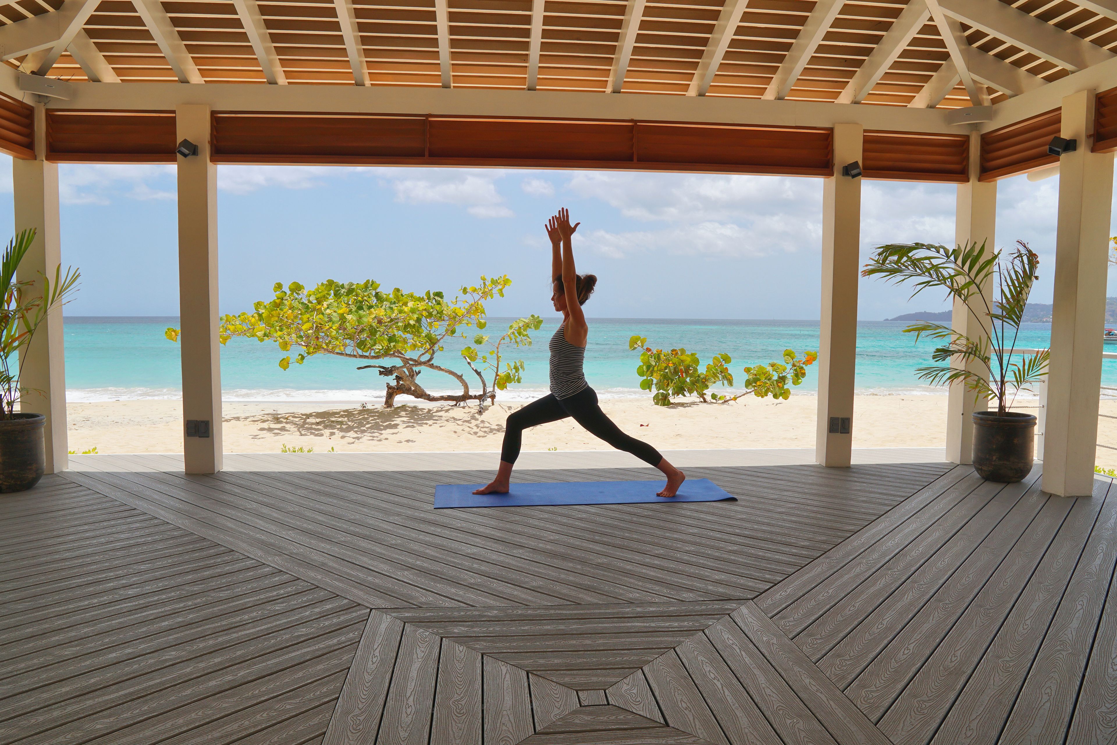 A wooden deck with a person performing a yoga pose, overlooking a beach with clear blue water and palm trees.