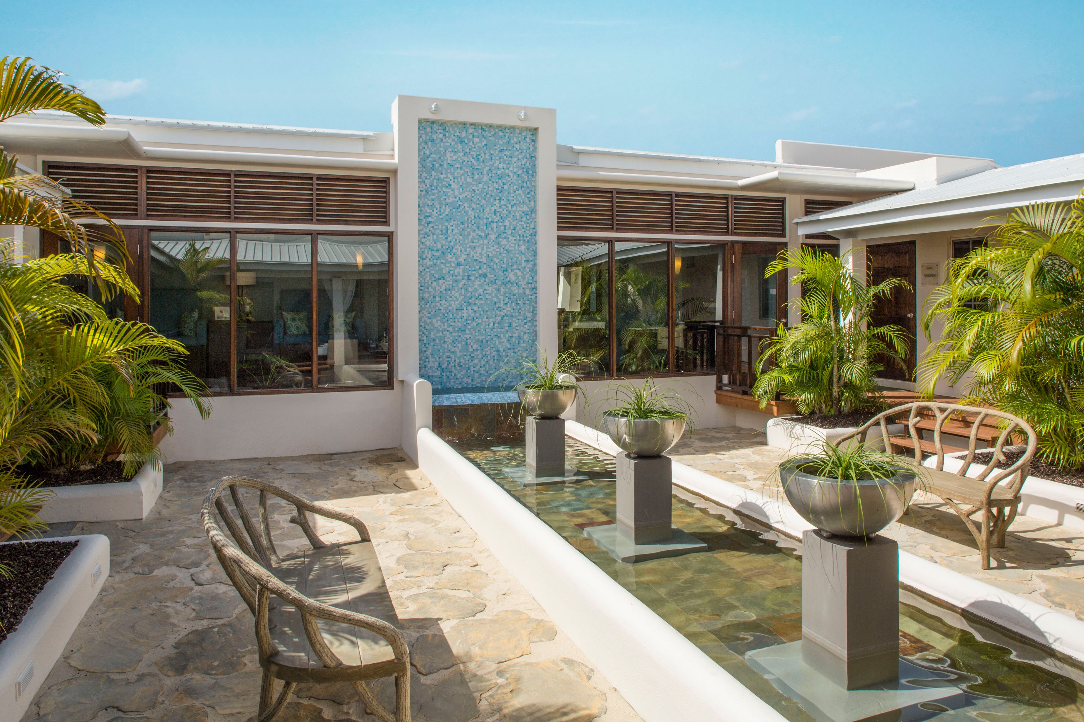 A modern outdoor area with stone flooring, a central water feature, and seating areas with planters.