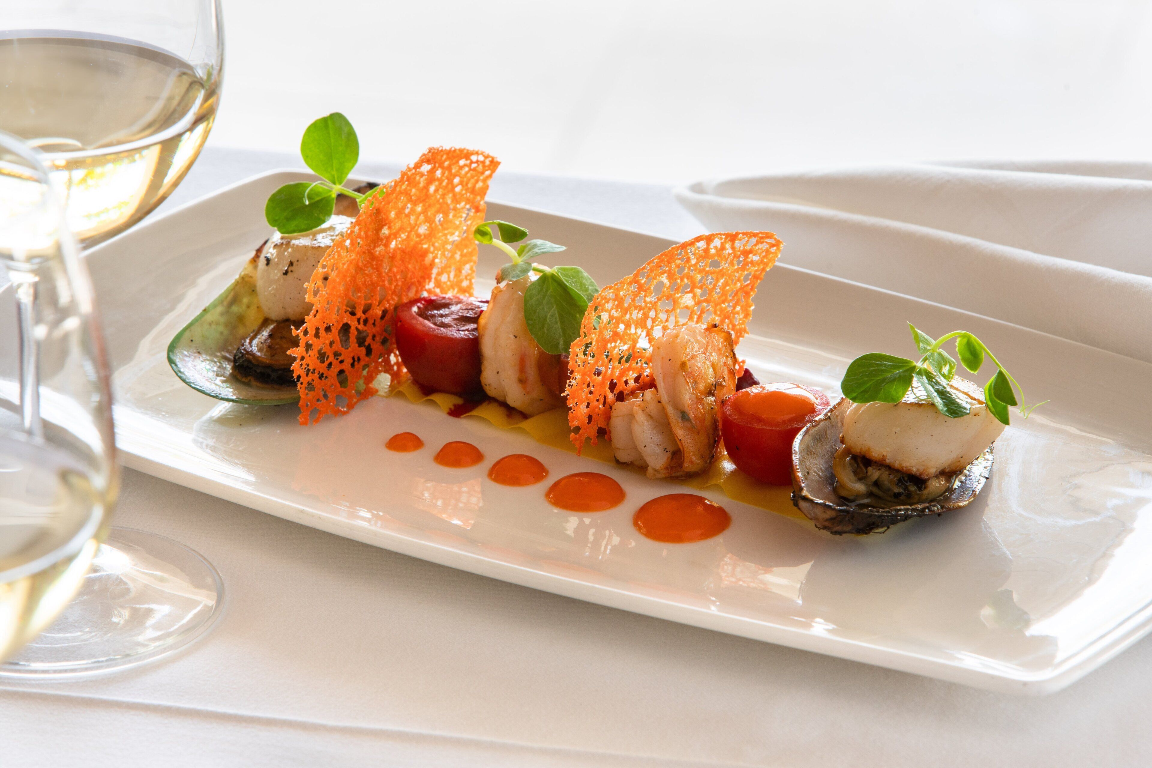A white rectangular plate with a seafood dish, garnished with orange netting and green leaves, accompanied by a glass of white wine.