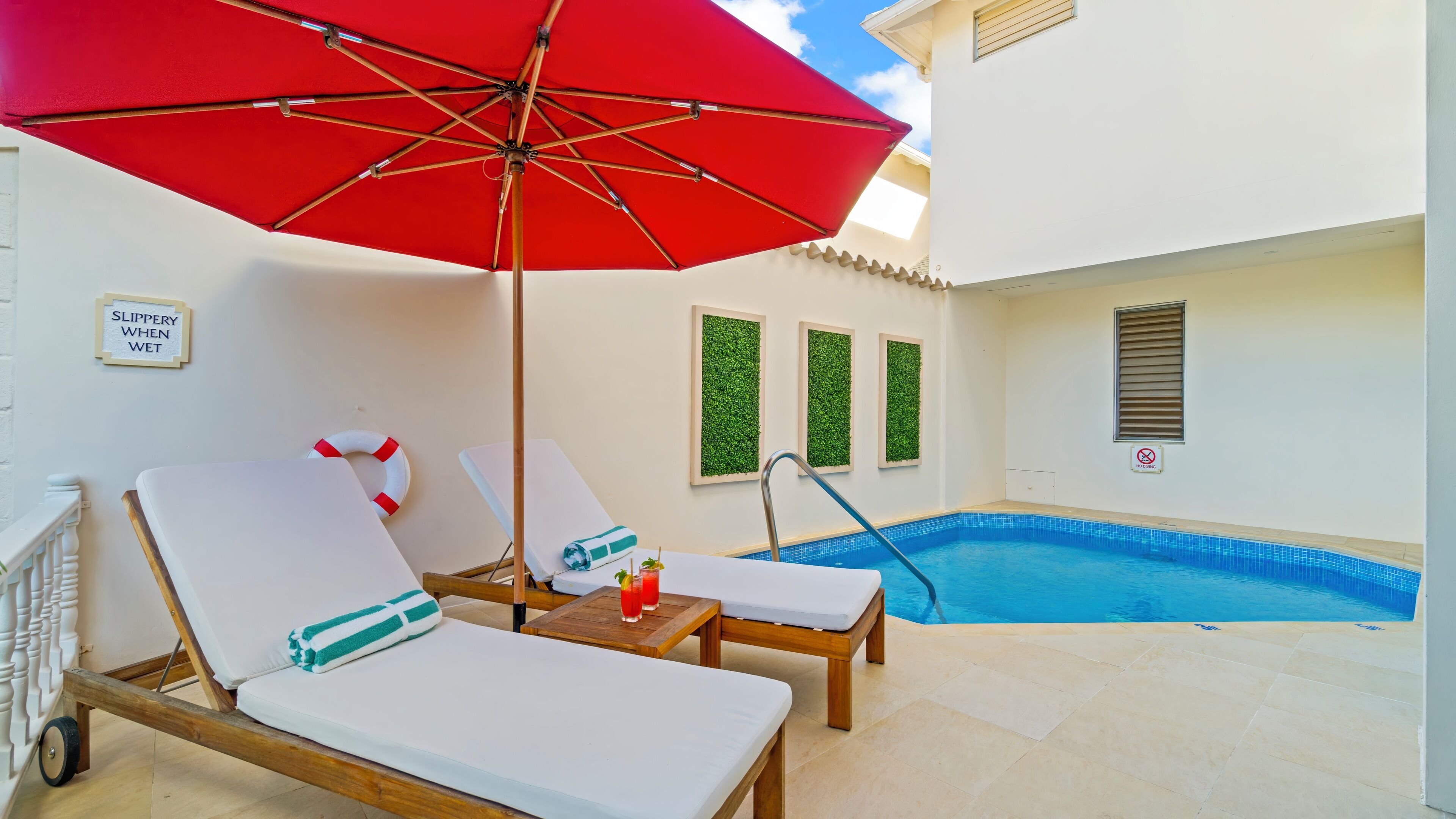 Zona de piscina con dos sillones reclinables, una sombrilla roja y un cartel que indica que la piscina es resbaladiza cuando está mojada.