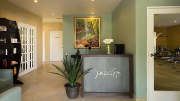 A reception area with a reception desk, a potted plant, a painting, and a door leading to a gym.