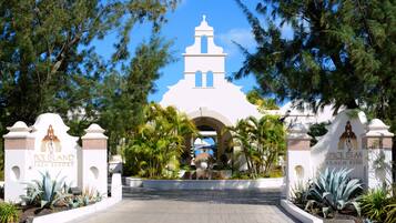 Entrada a un complejo turístico con un edificio blanco, palmeras y una fuente.