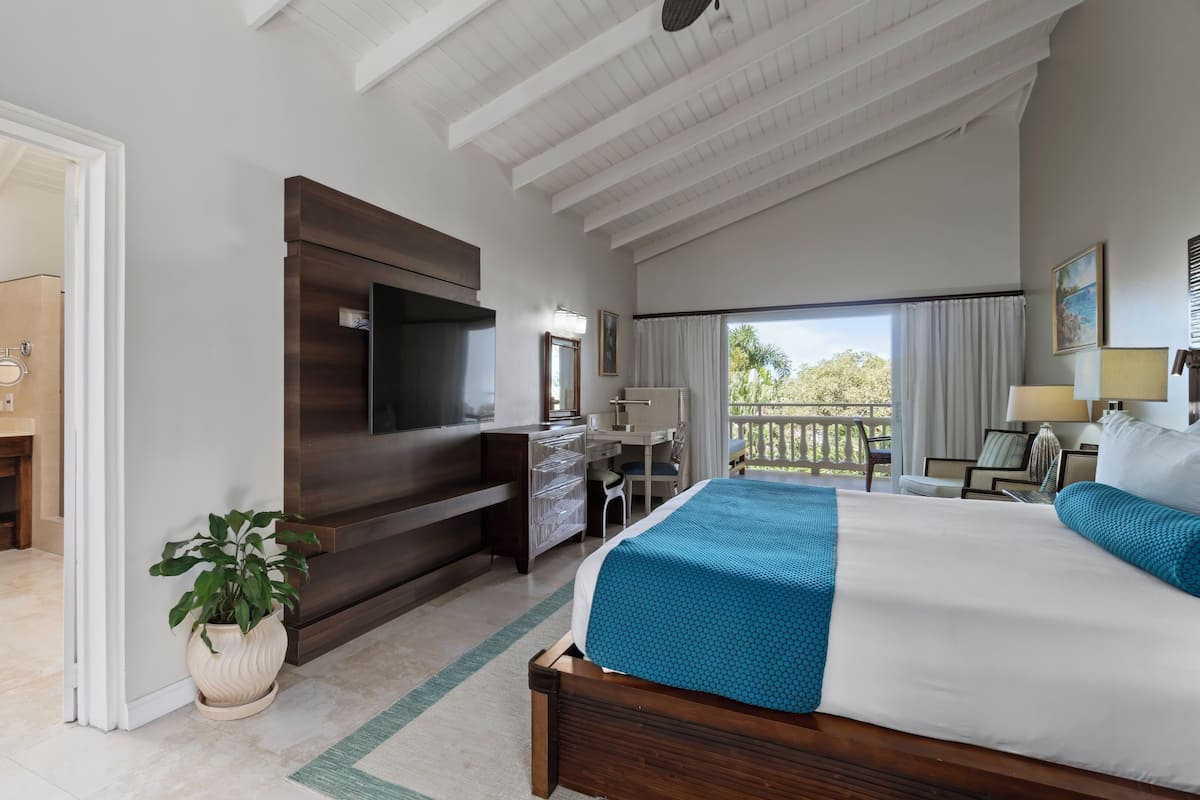 A hotel room with a large bed, a desk, a TV, a balcony with a view, and a bathroom with a mirror and sink.