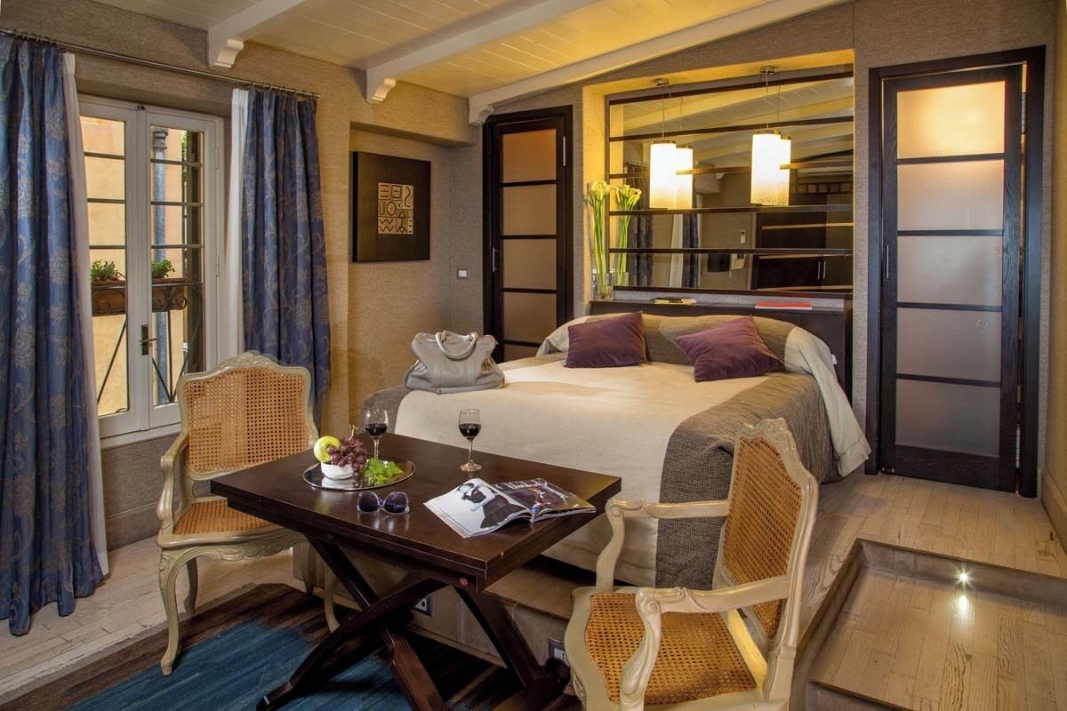 A hotel room with a bed, two chairs, a table with a magazine and wine glasses, and a window with curtains.