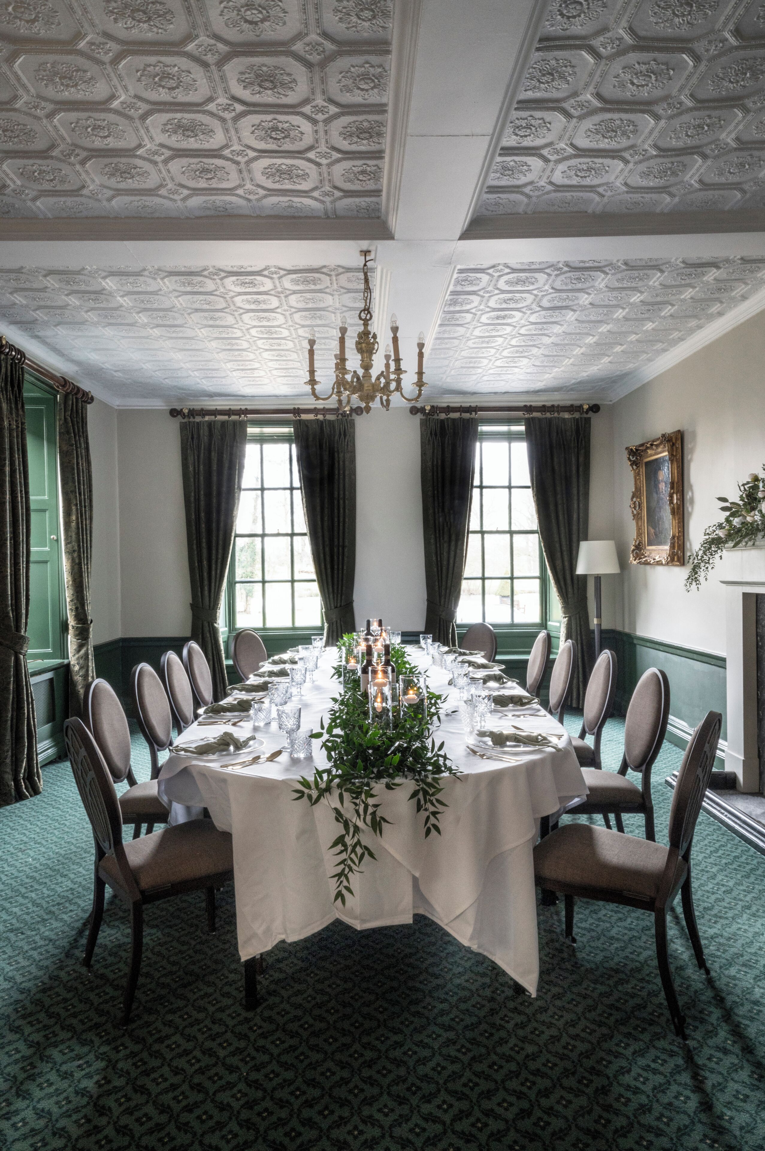 A formal dining room with a round table set for a meal, surrounded by chairs, with a chandelier and a window with curtains.