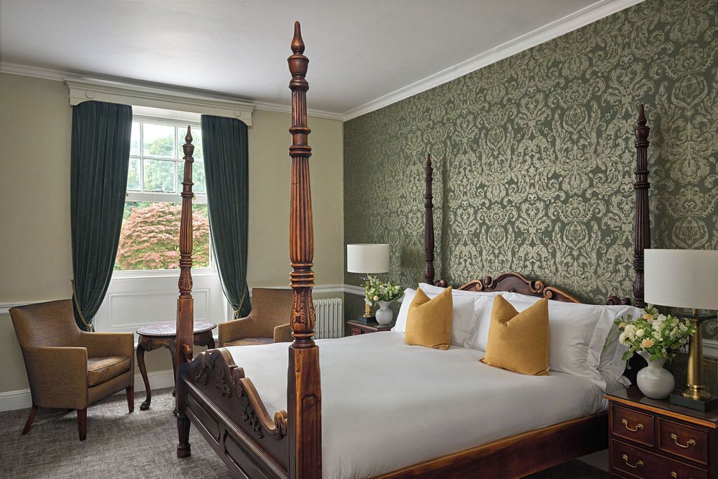 A four-poster bed with a wooden headboard, a chair, a nightstand with a lamp, and a window with curtains.