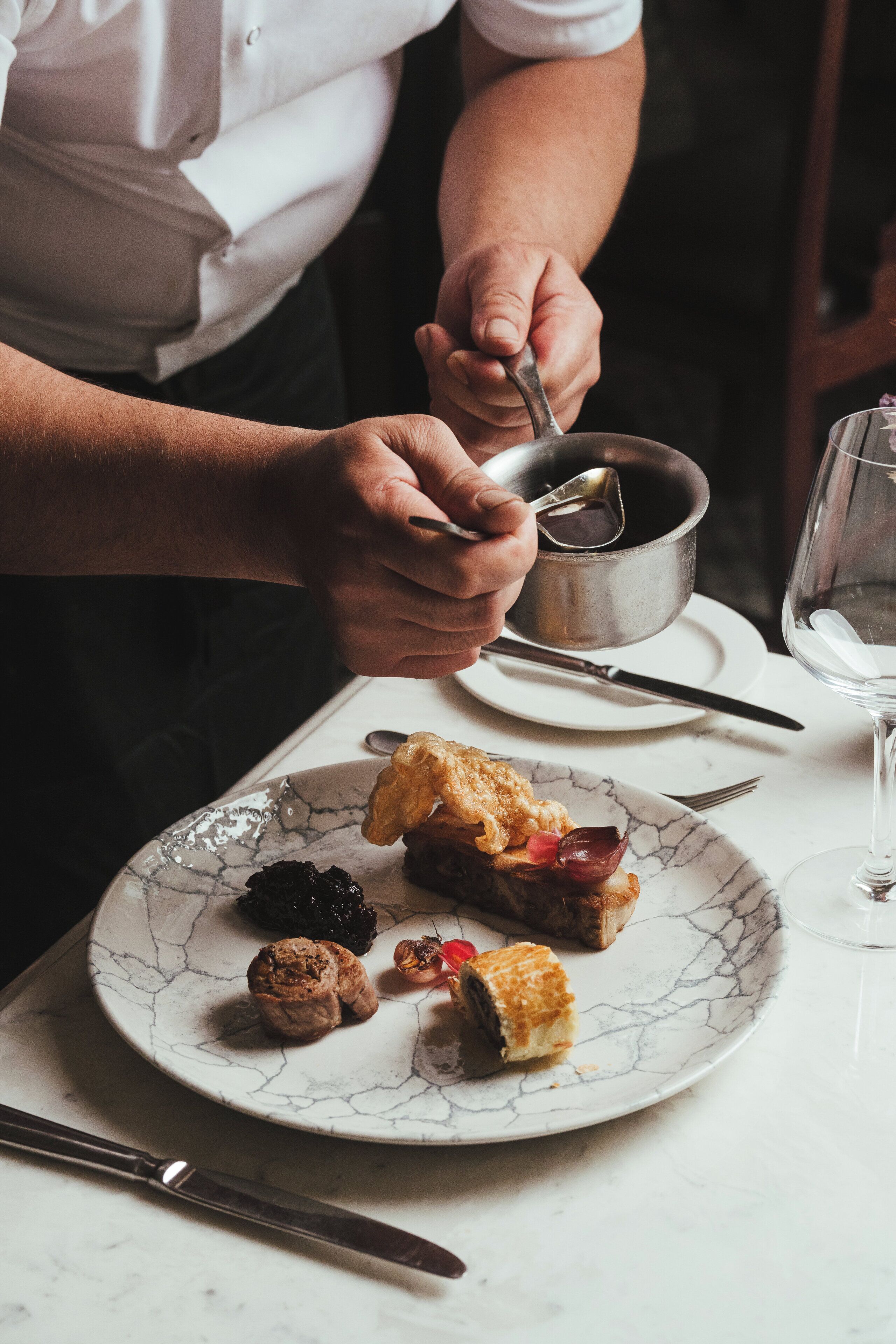 A person serving food on a plate with a spoon, a wine glass, and a knife on the table.