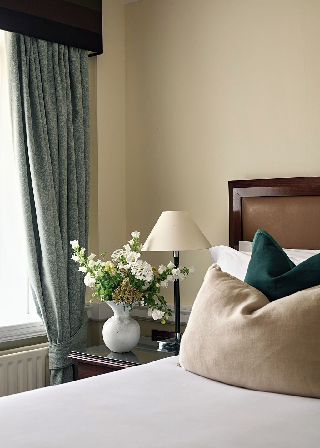 Habitación de hotel con cama, mesita de noche con lámpara y un jarrón con flores, y una ventana con cortinas.