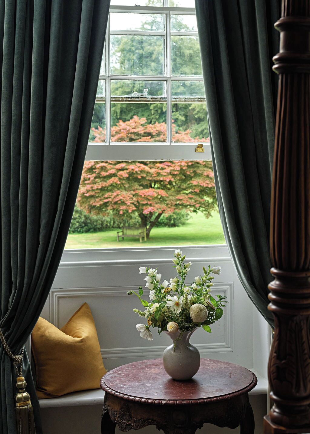A room with a window, a vase with flowers, a round wooden table, and a yellow pillow.