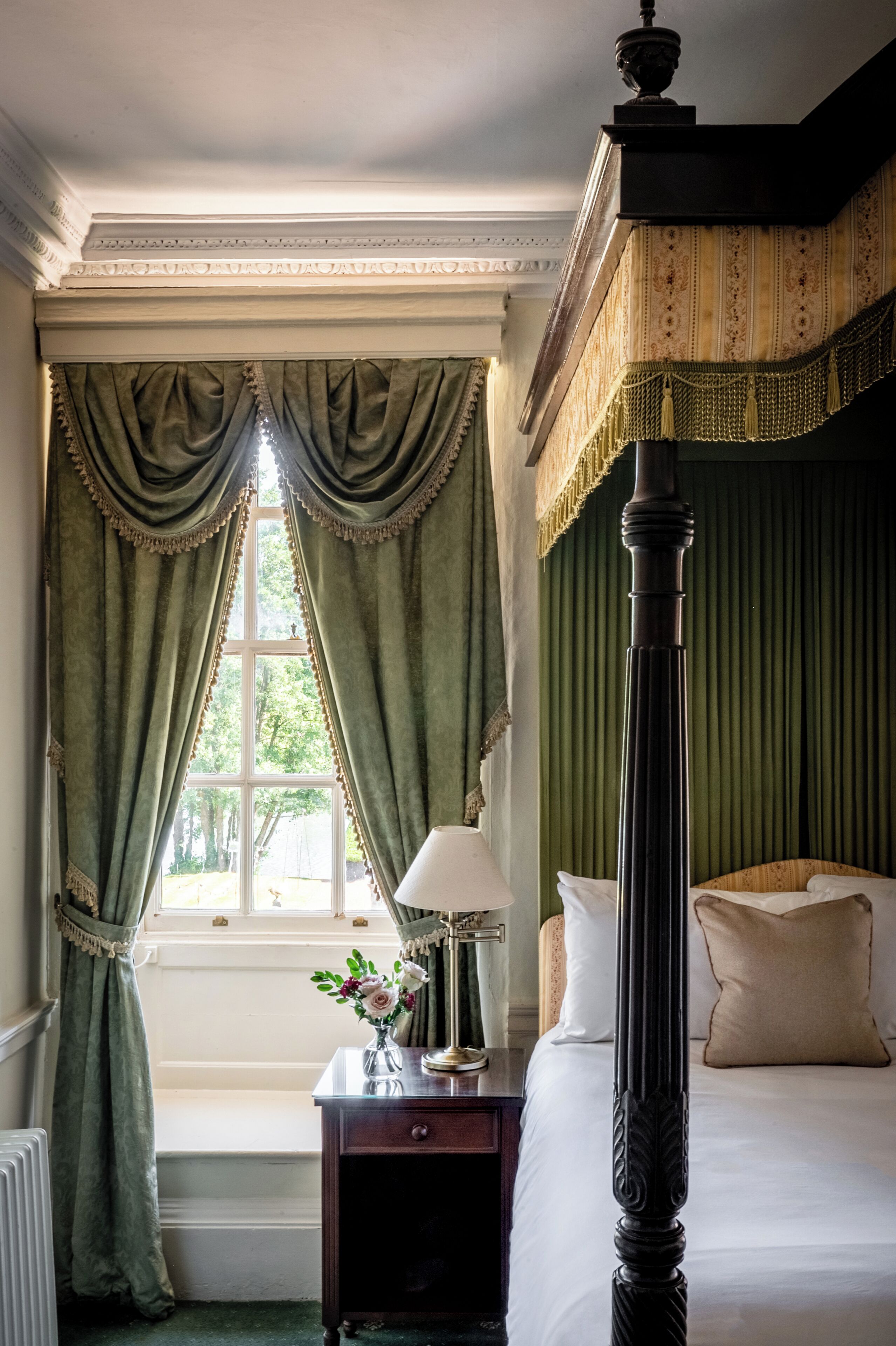 A room with a four-poster bed, a nightstand with a lamp, and a window with curtains.
