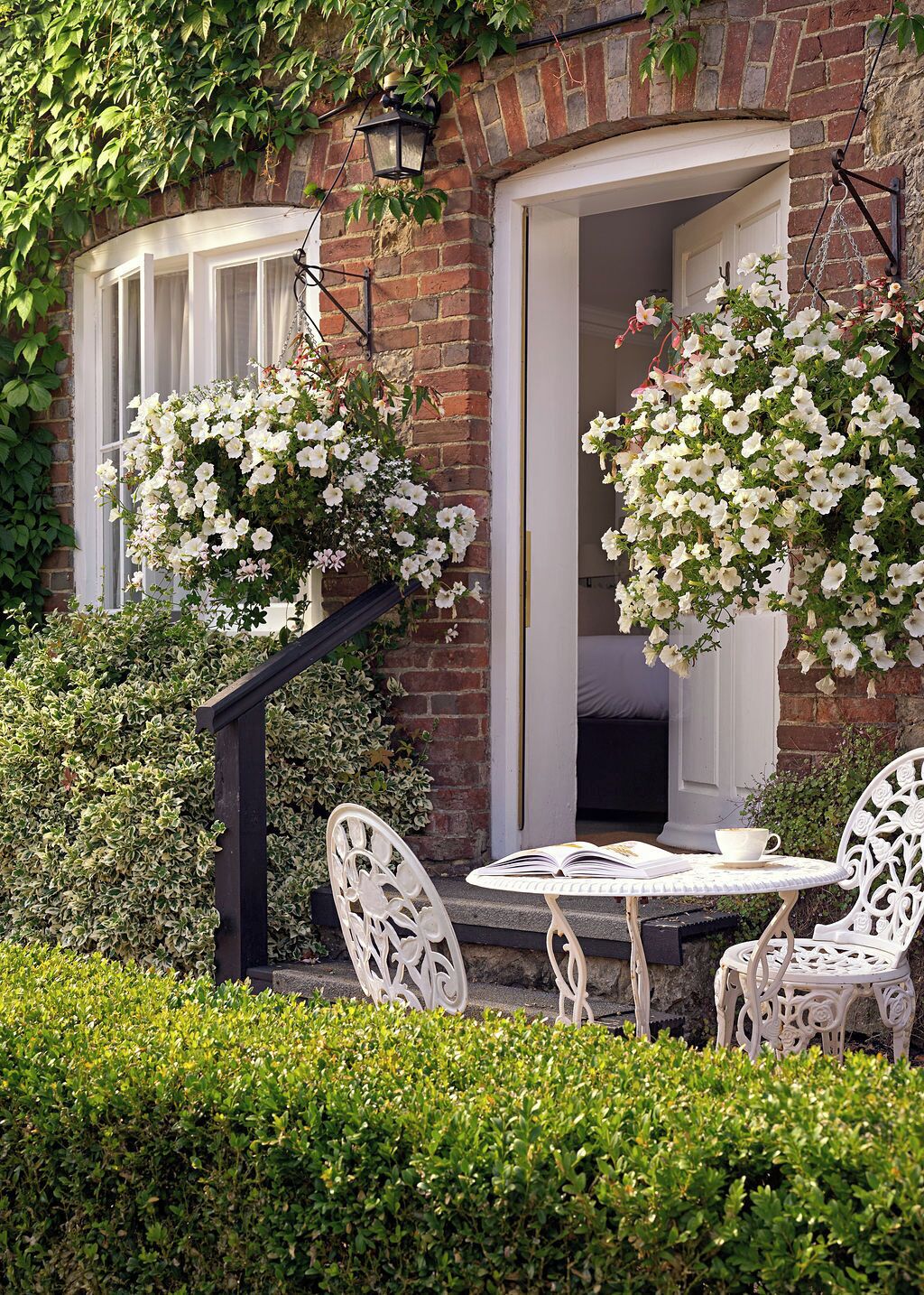 A brick building with white-framed windows, a hanging flower basket, and a table with chairs in front.