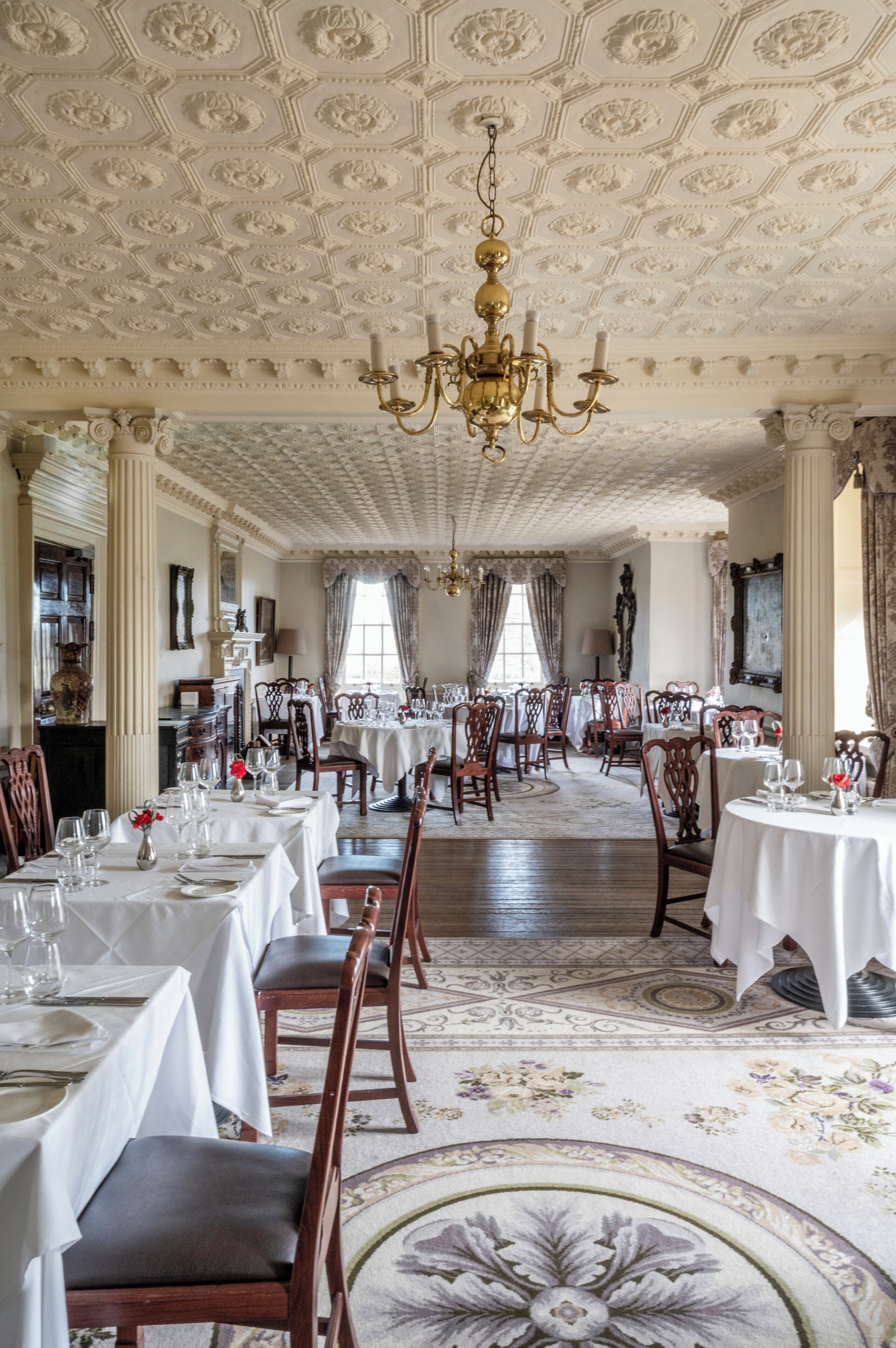 A formal dining room with multiple tables set for service, ornate ceiling decorations, and classical architecture.