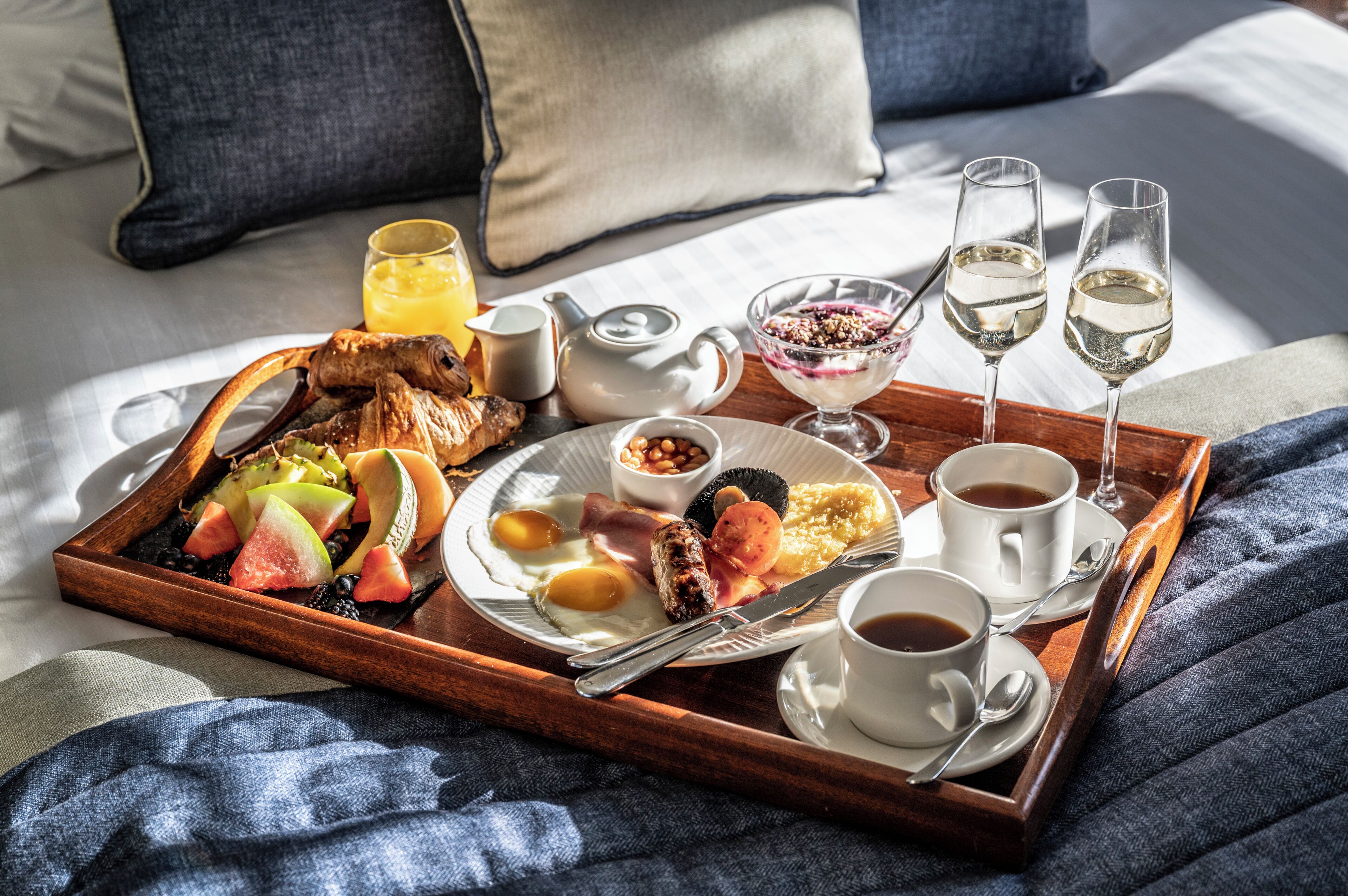 Ein Frühstücksbrett mit Eiern, Würstchen, Toast und Obst auf einem Bett mit Kissen und Decke.