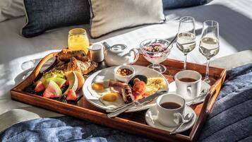 Ein Frühstücksbrett mit Eiern, Würstchen, Toast und Obst auf einem Bett mit Kissen und Decke.