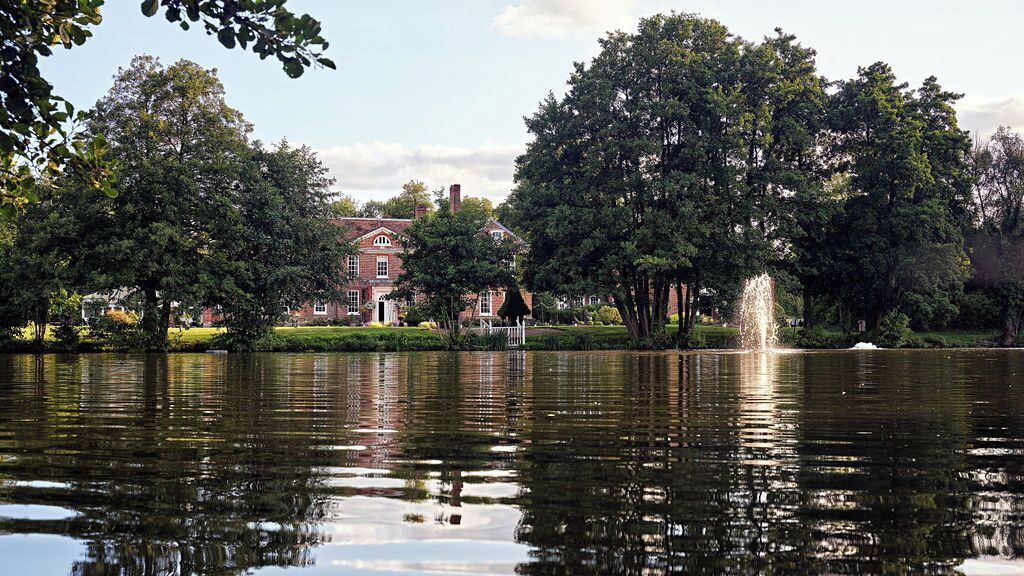 A calm lake with a fountain, surrounded by lush trees and a red brick building in the background.