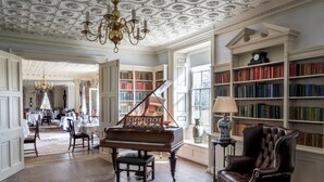 A grand room with a grand piano, a leather armchair, and a bookshelf filled with books. - Chilston Park Hotel (Maidstone)