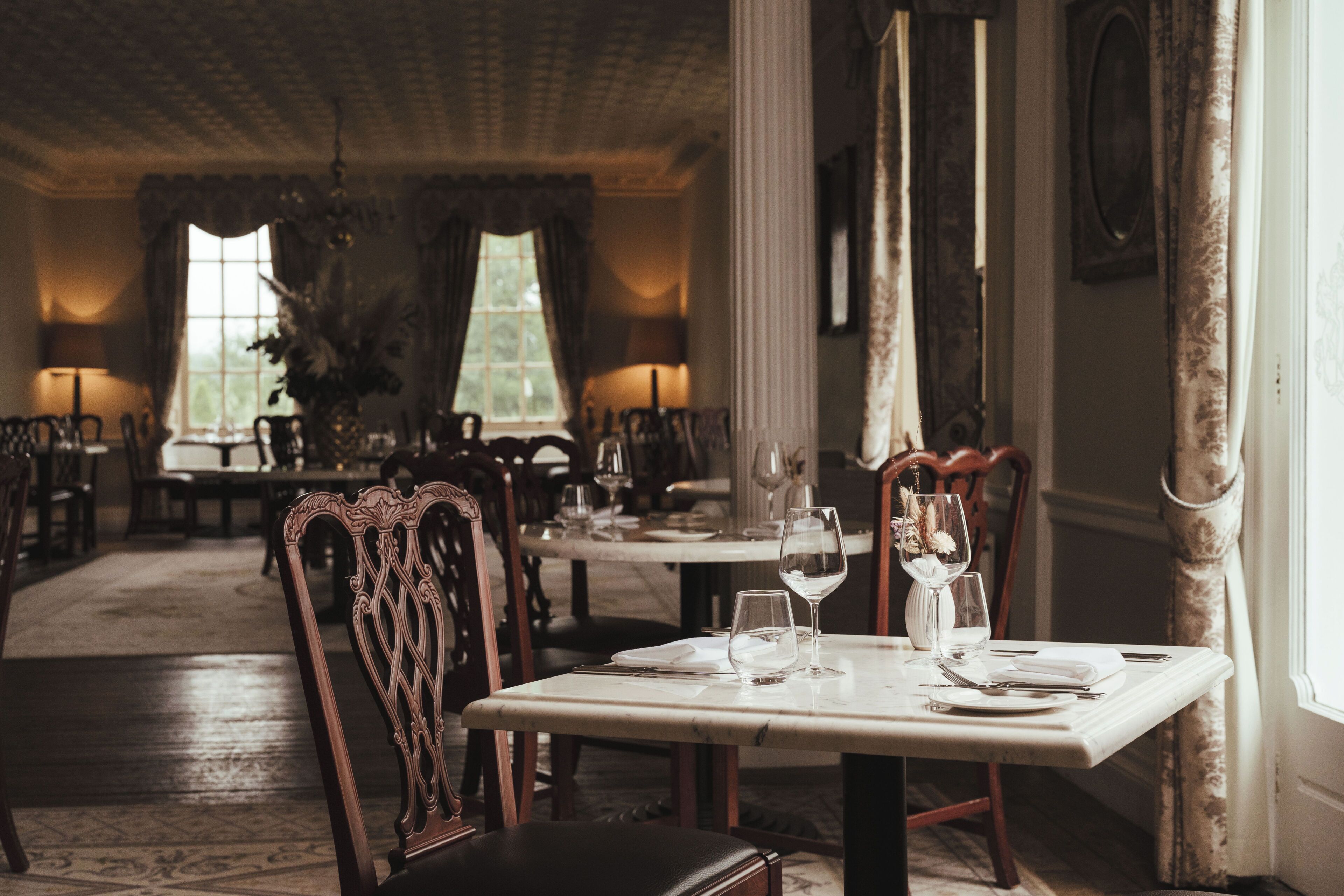 A dining area with tables set for two, chairs, and a vase with dried flowers.