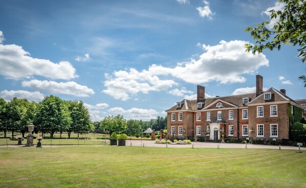 Stately brick building with a central entrance and two wings, set amidst a serene lawn and lush trees.