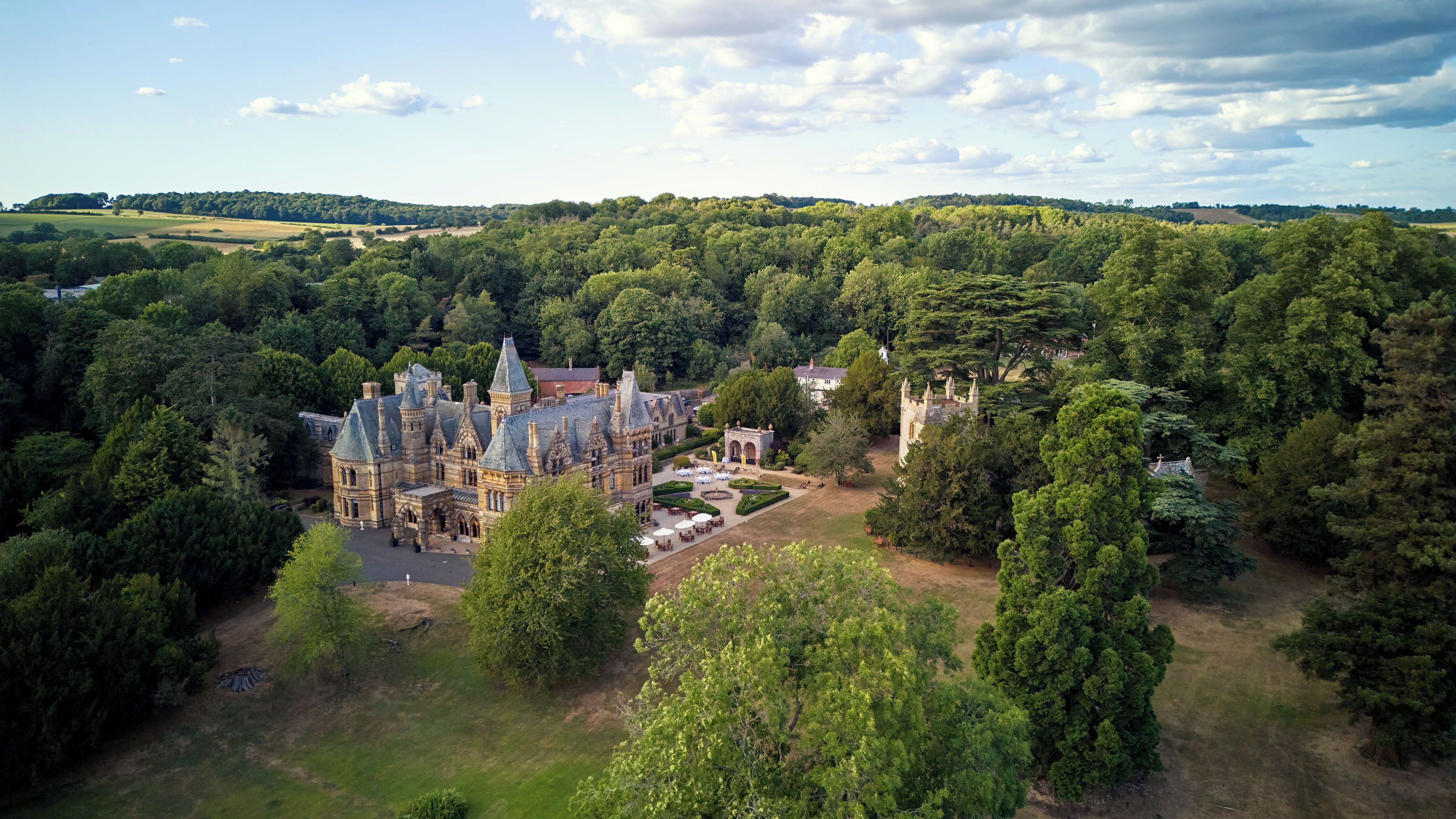 A large, historic building surrounded by dense forest, with a formal garden and a small castle-like structure nearby.