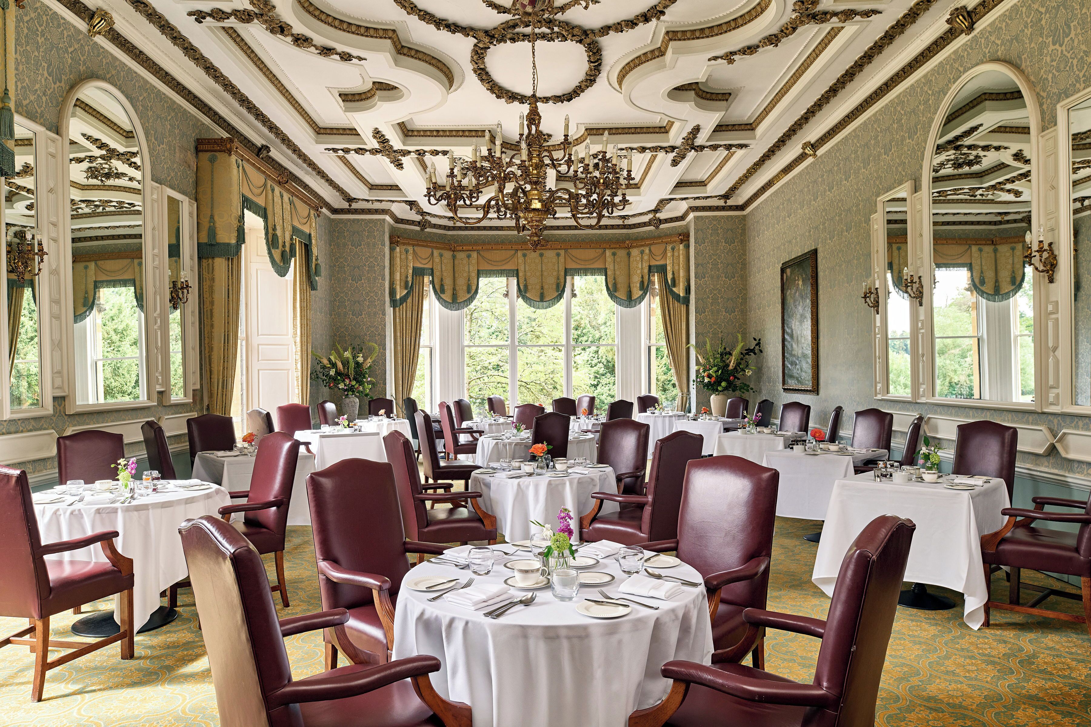 Uma sala de jantar formal com várias mesas e cadeiras, decoração de teto elaborada e amplas janelas que permitem a entrada de luz natural.