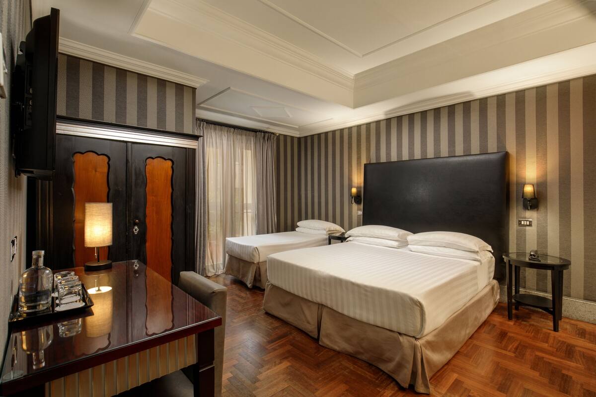 A hotel room with two beds, a desk with a lamp, a minibar, and a large mirror.