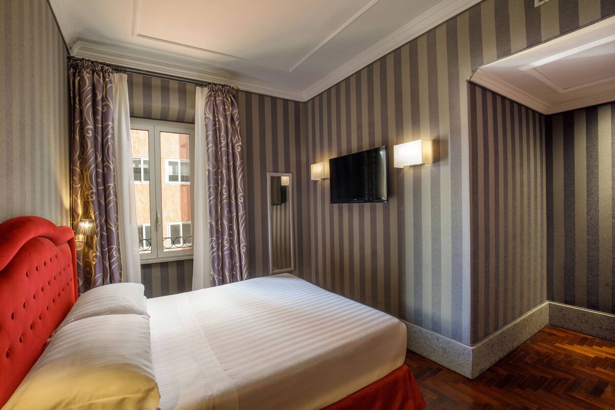 A hotel room with a bed, striped wallpaper, a TV, and a window with curtains.