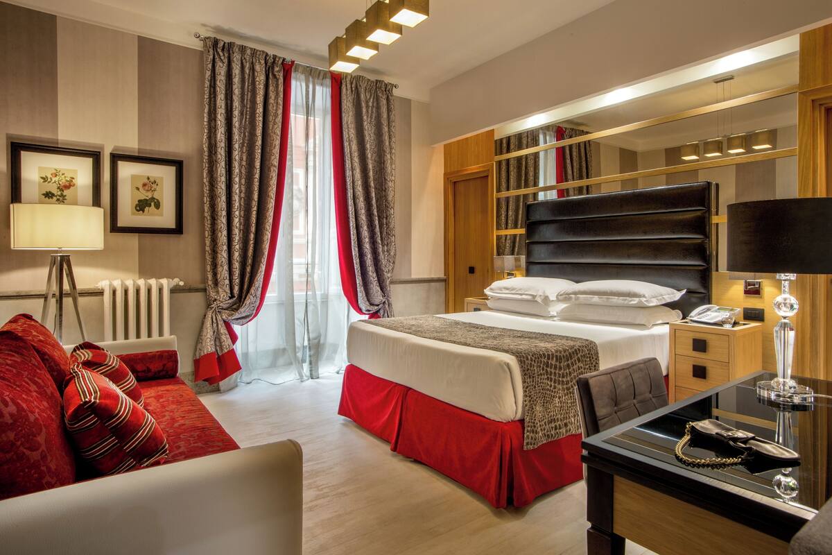 A hotel room with a bed, a desk, a chair, a red sofa, a lamp, a mirror, a wardrobe, and a window with curtains.