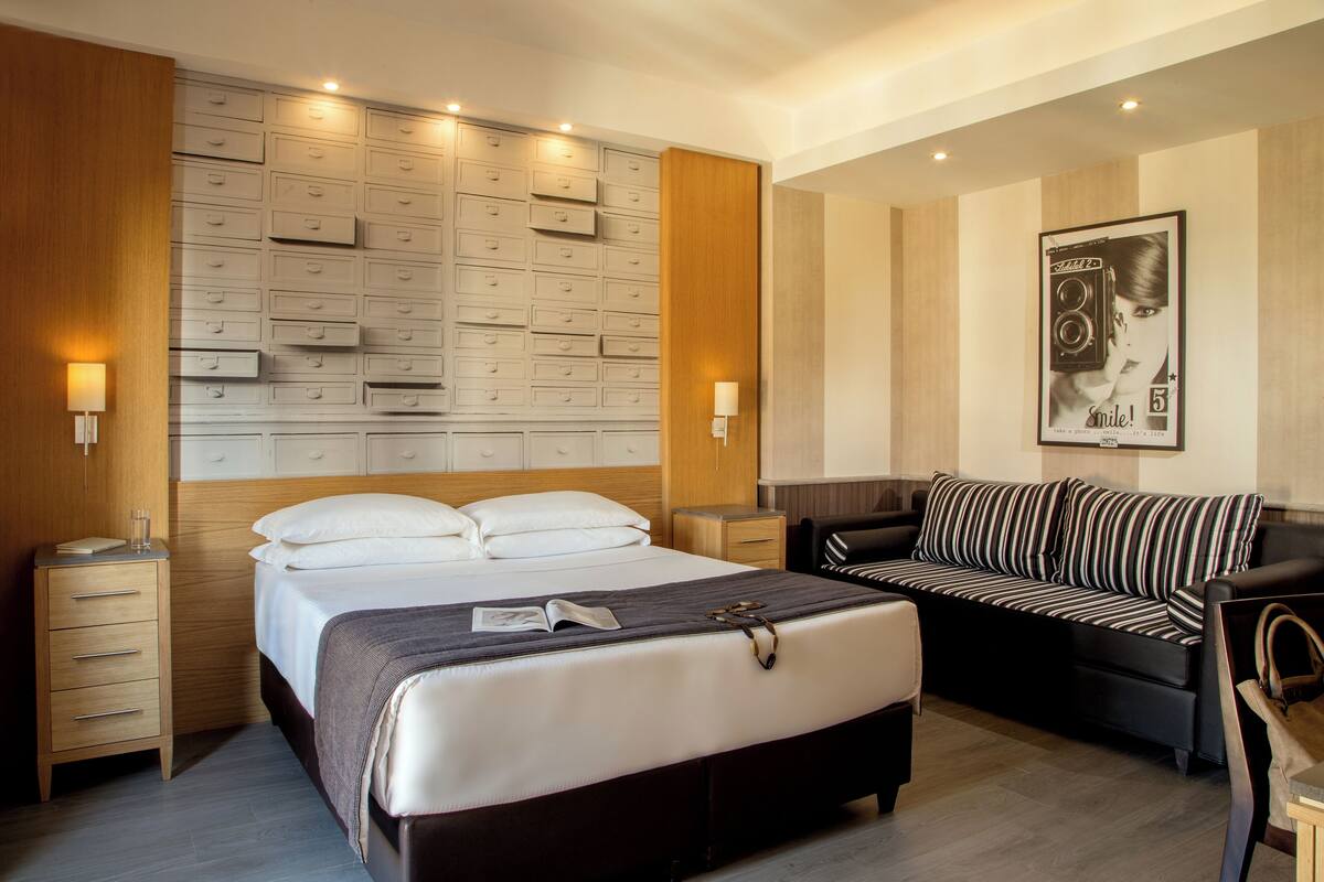 A modern hotel room with a large bed, a sofa, a nightstand, and a wall unit with drawers.