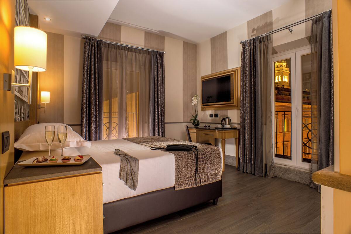 A hotel room with a bed, a desk, a TV, and a view of a building through the window.