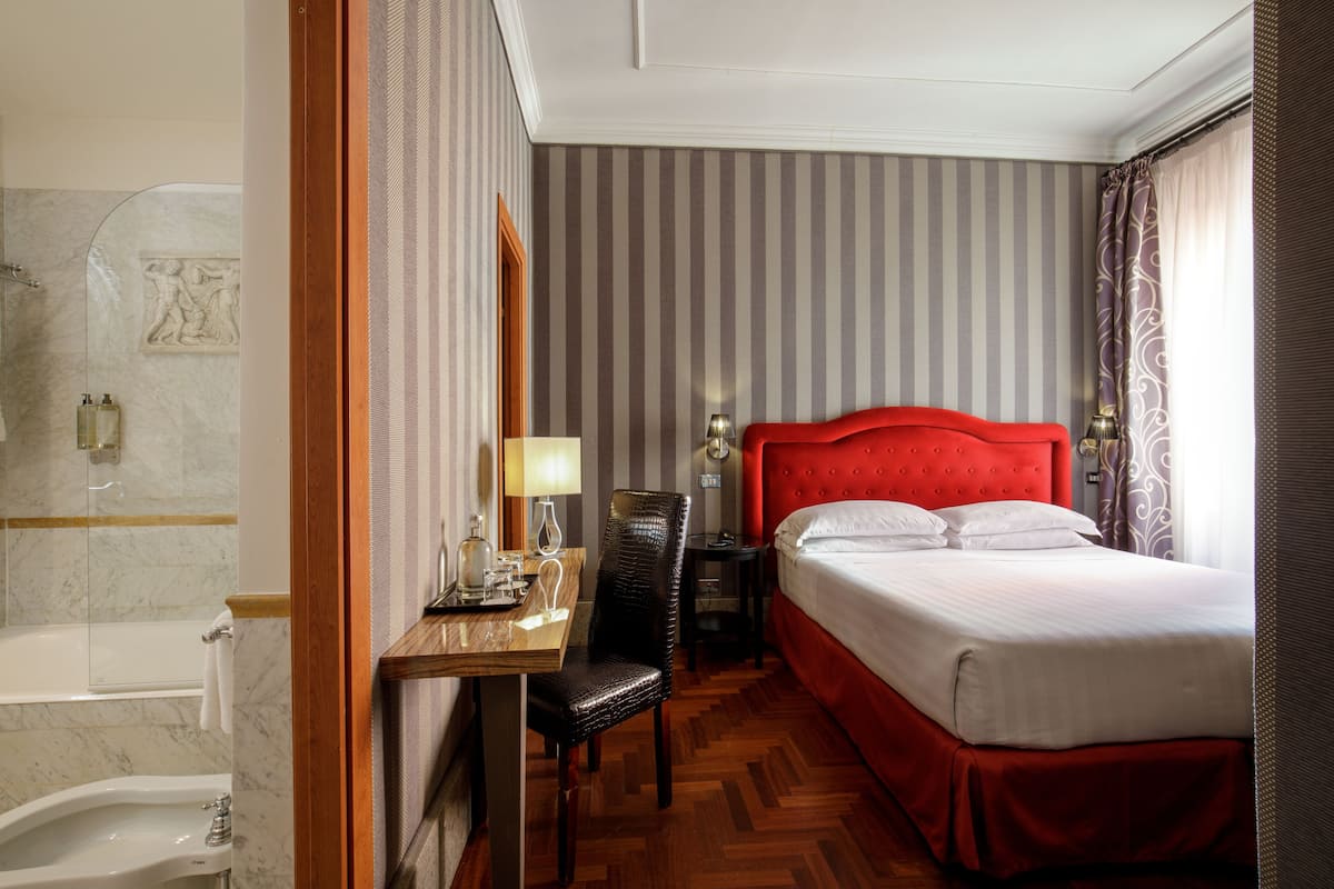 A hotel room with a red upholstered headboard, a wooden desk with a chair, a bedside table with a lamp, and a large bed with white linens.