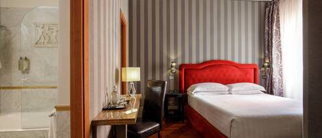 A hotel room with a red upholstered headboard, a wooden desk with a chair, a bedside table with a lamp, and a large bed with white linens.