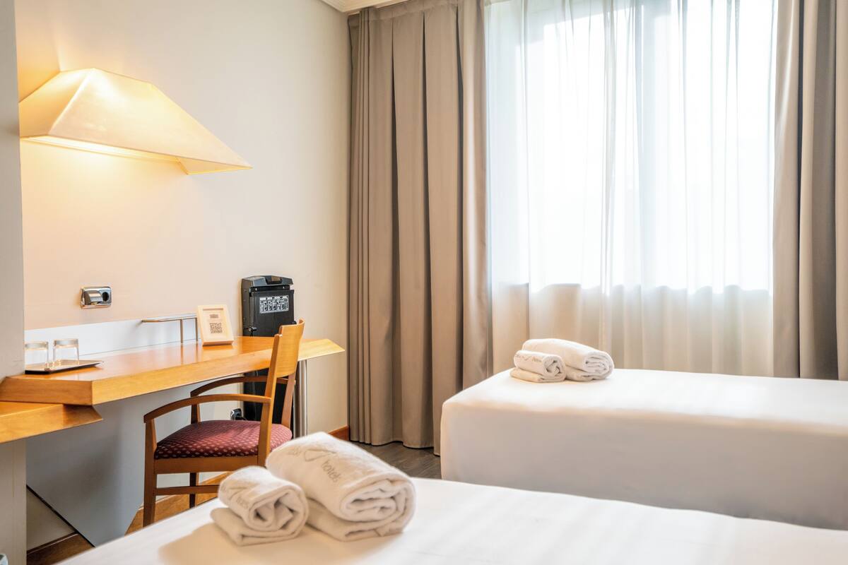 A hotel room with a desk, two beds, a chair, a lamp, and a window with curtains.