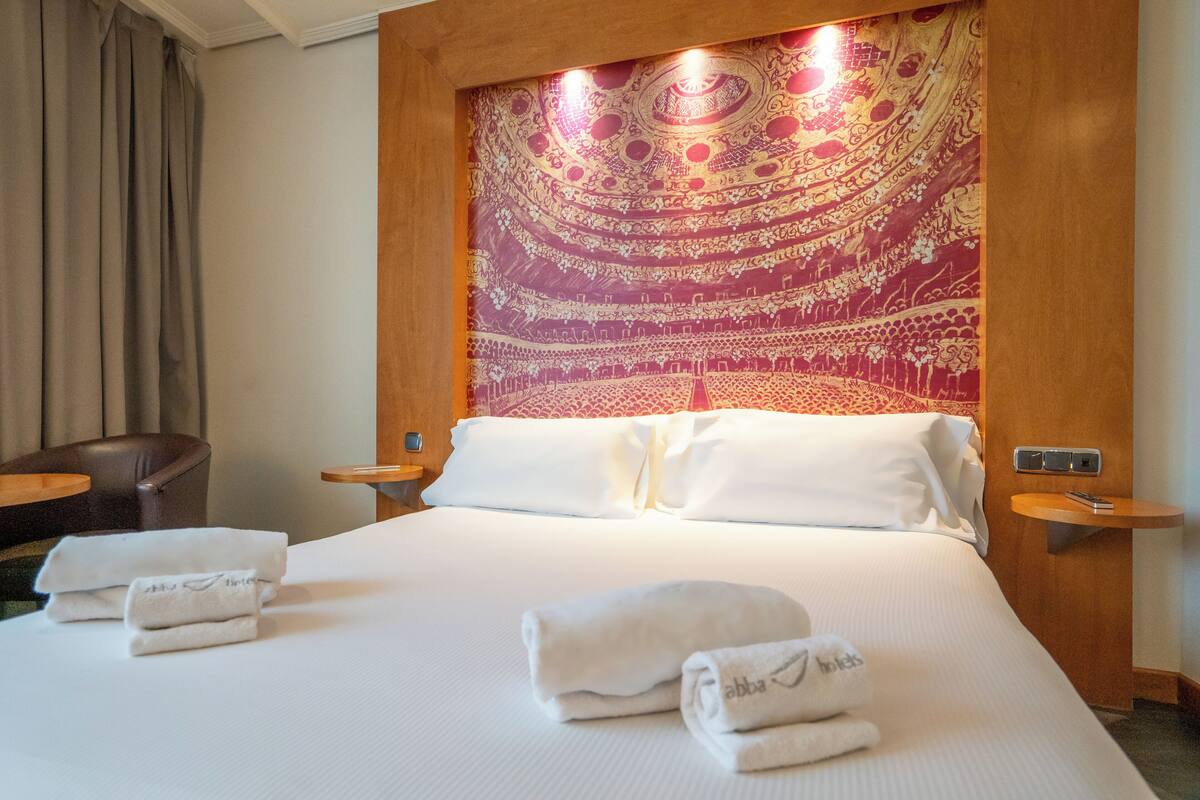 A hotel room with a large bed, white linens, a tapestry headboard, and two bedside tables with lamps.