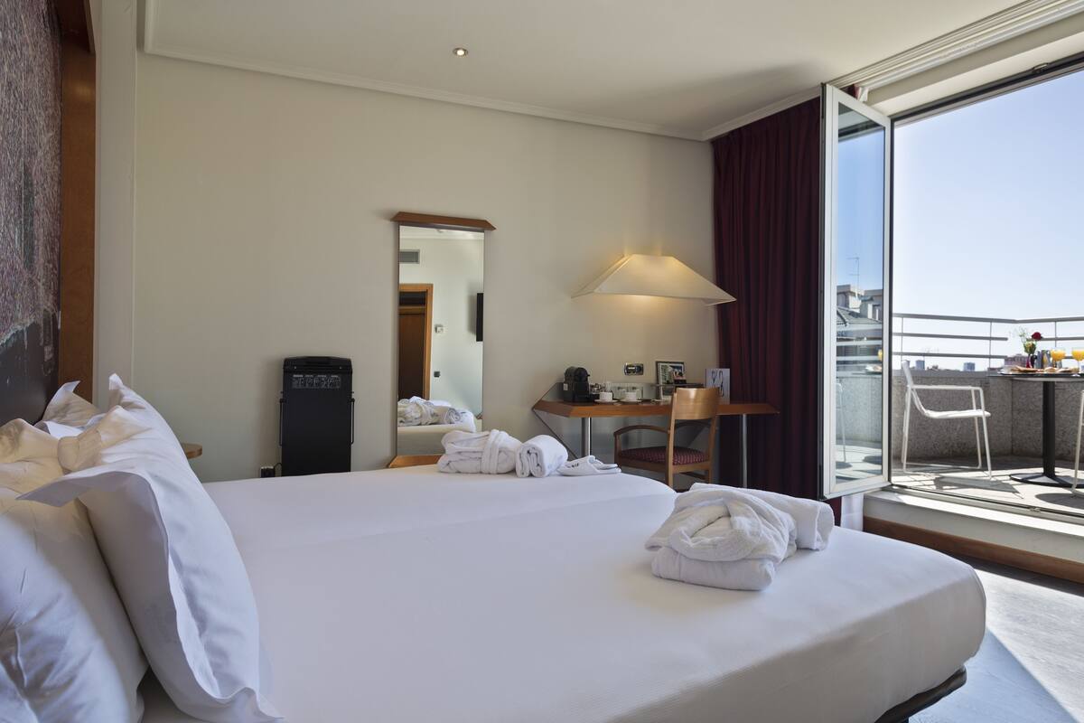 A hotel room with a large bed, a desk with a chair, a mirror, a lamp, and a view of the city through a large window.