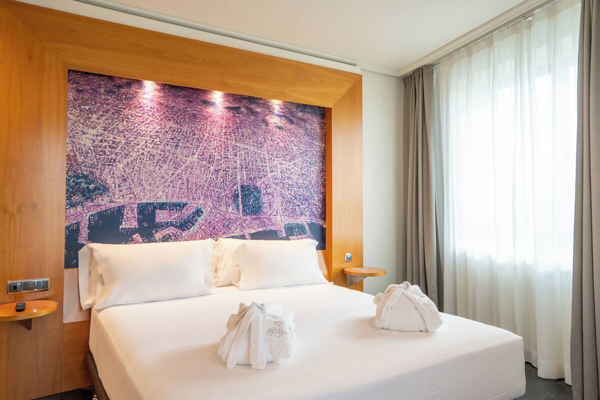 A hotel room with a large bed, two pillows, a wooden headboard, a nightstand, and a window with curtains.