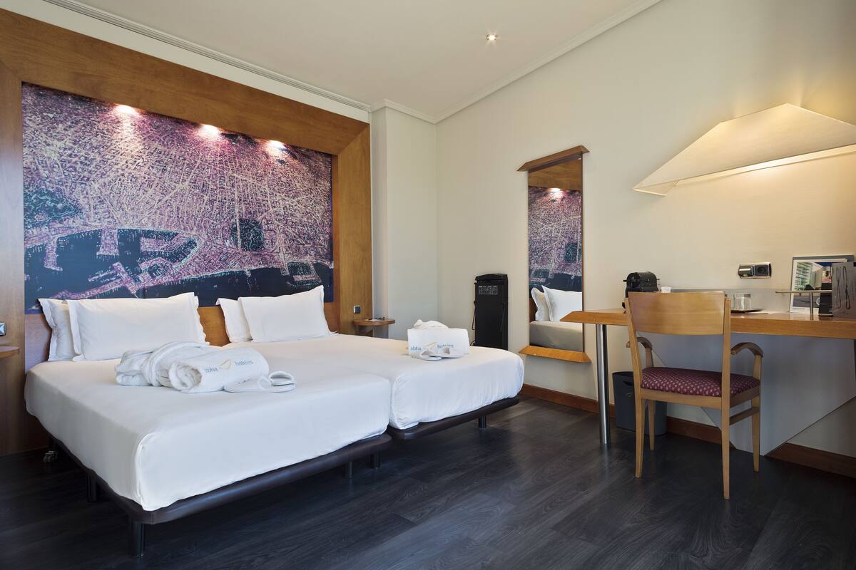 A hotel room with a large bed, two bedside tables, a desk with a chair, a large mirror, and a cityscape artwork on the wall.