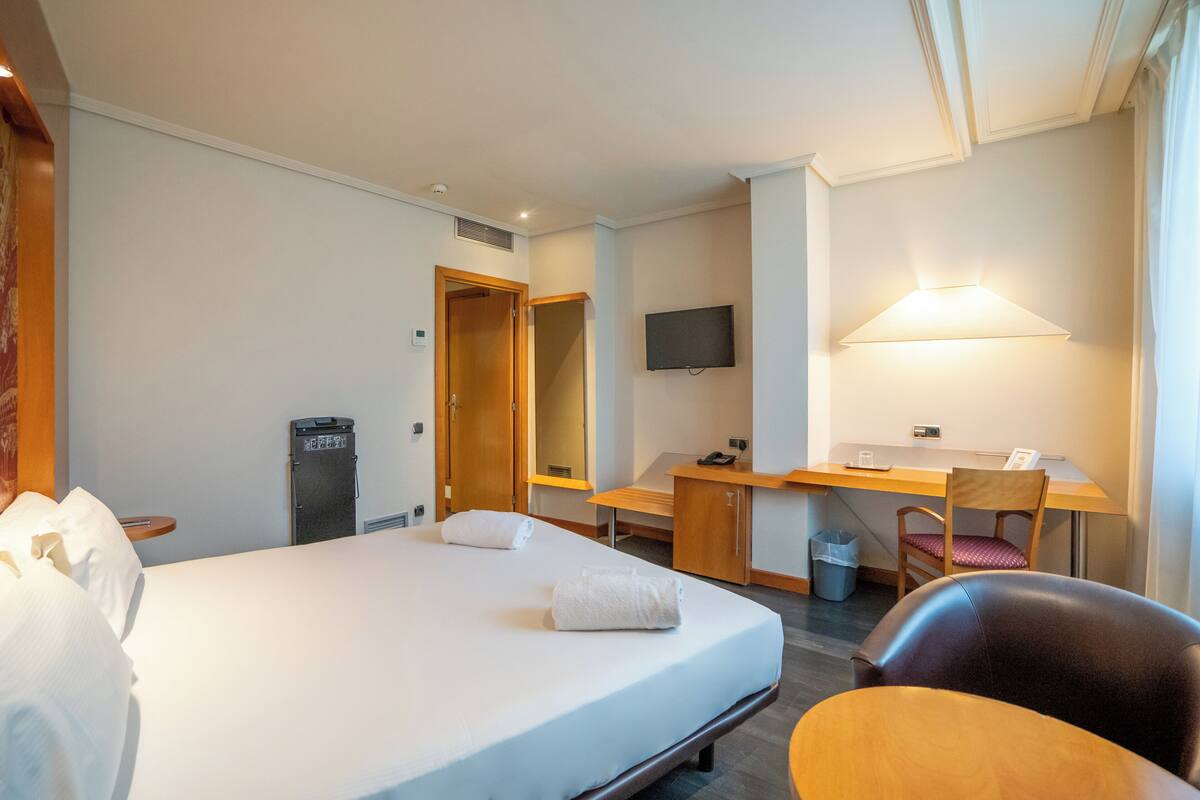 A hotel room with a bed, a desk, a chair, a TV, and a lamp.
