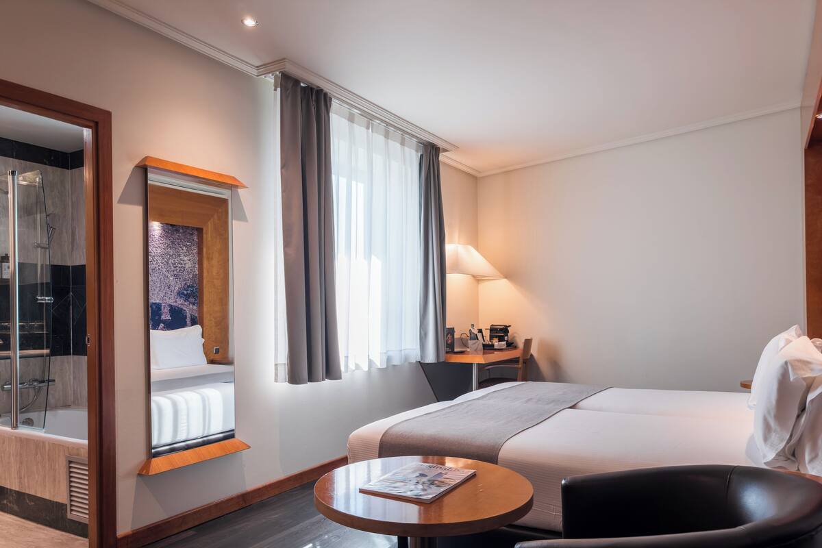 A hotel room with a bed, a desk with a lamp, a chair, a round table, and a shower area.