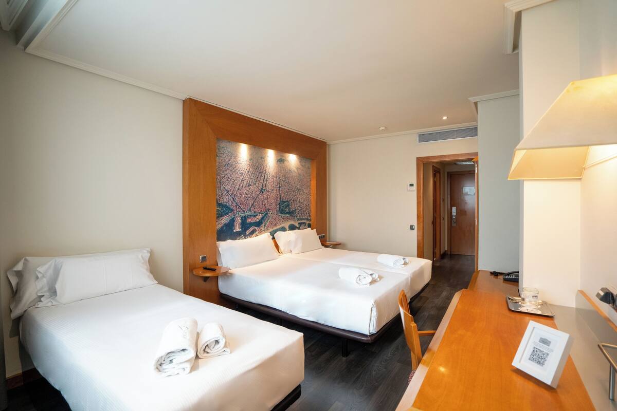 A hotel room with two beds, a wooden headboard, a wall mural, a desk with a lamp, and a door leading to another room.