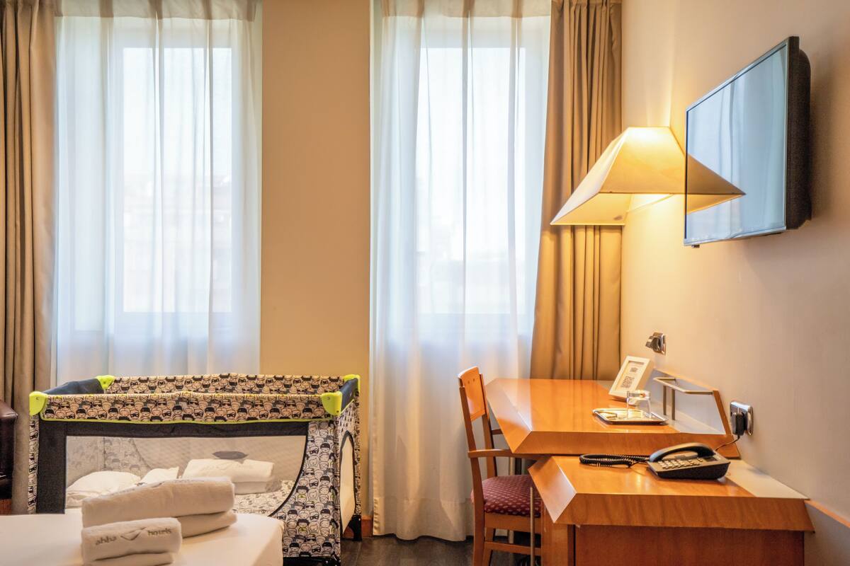 A hotel room with a bed, a desk with a lamp, a telephone, and a television mounted on the wall.