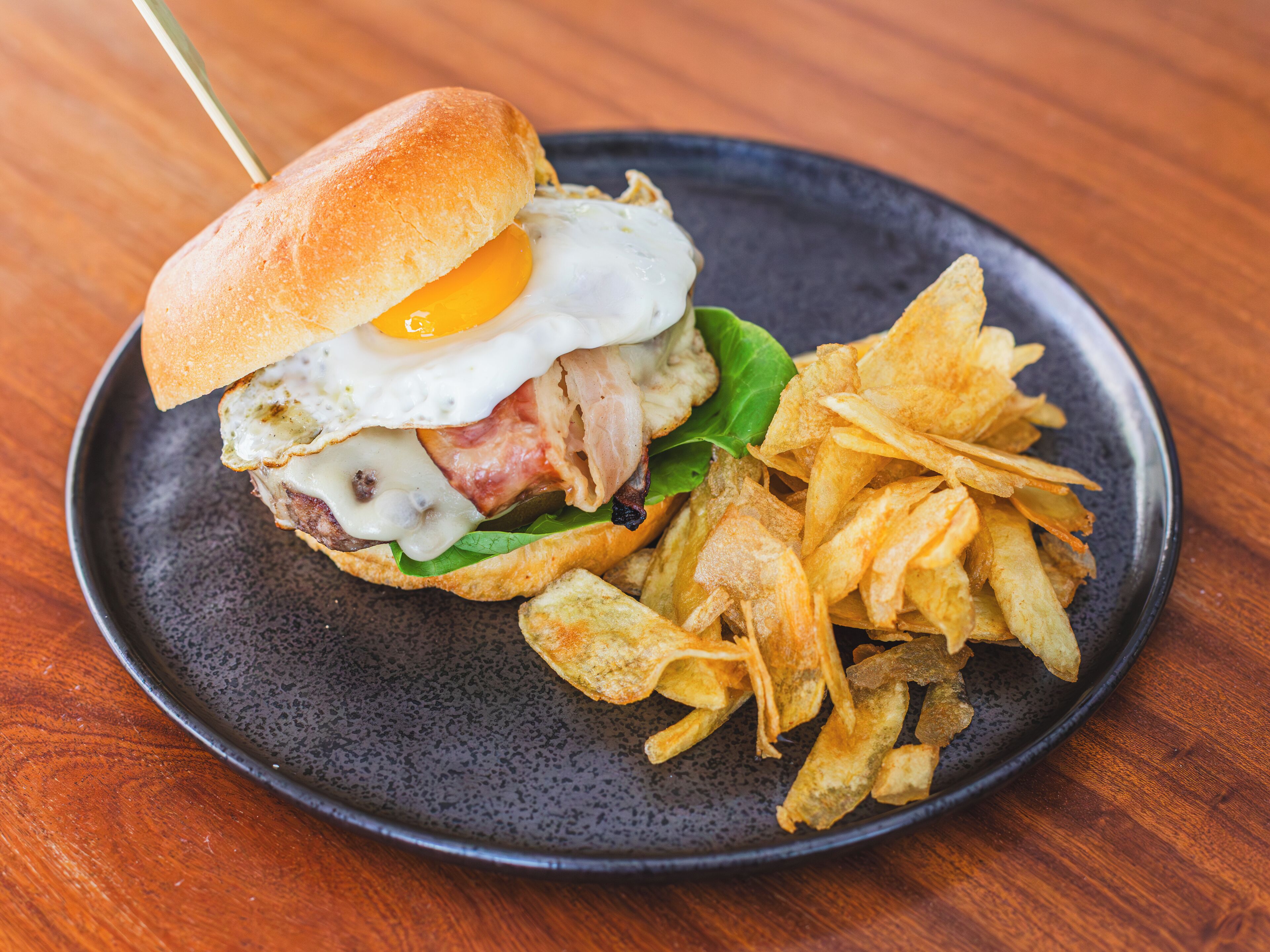 Un sándwich con huevo frito, tocino y queso en un pan, acompañado de una porción de papas fritas en un plato oscuro.