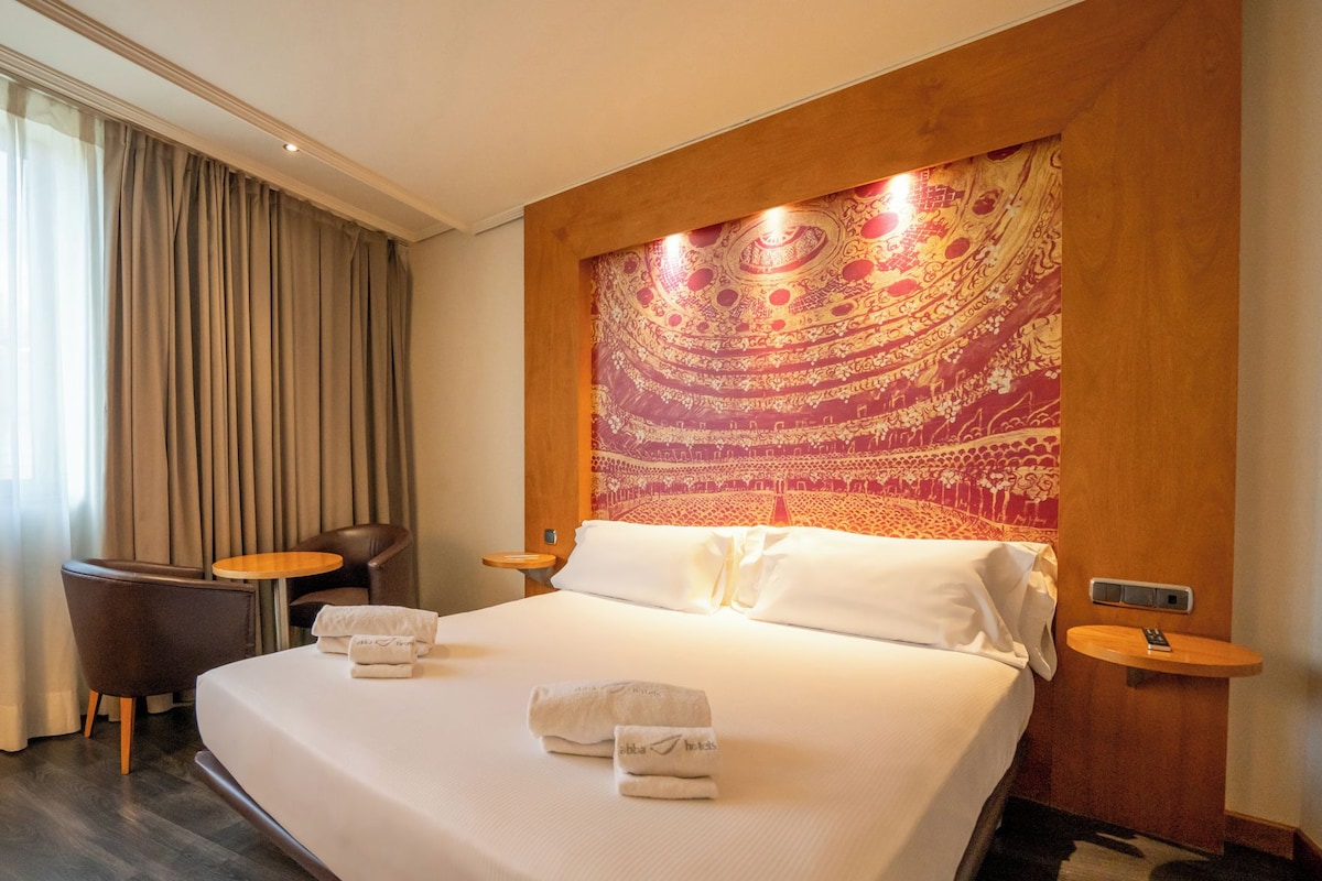 A hotel room with a large bed, two bedside tables, a chair, and a tapestry on the wall.