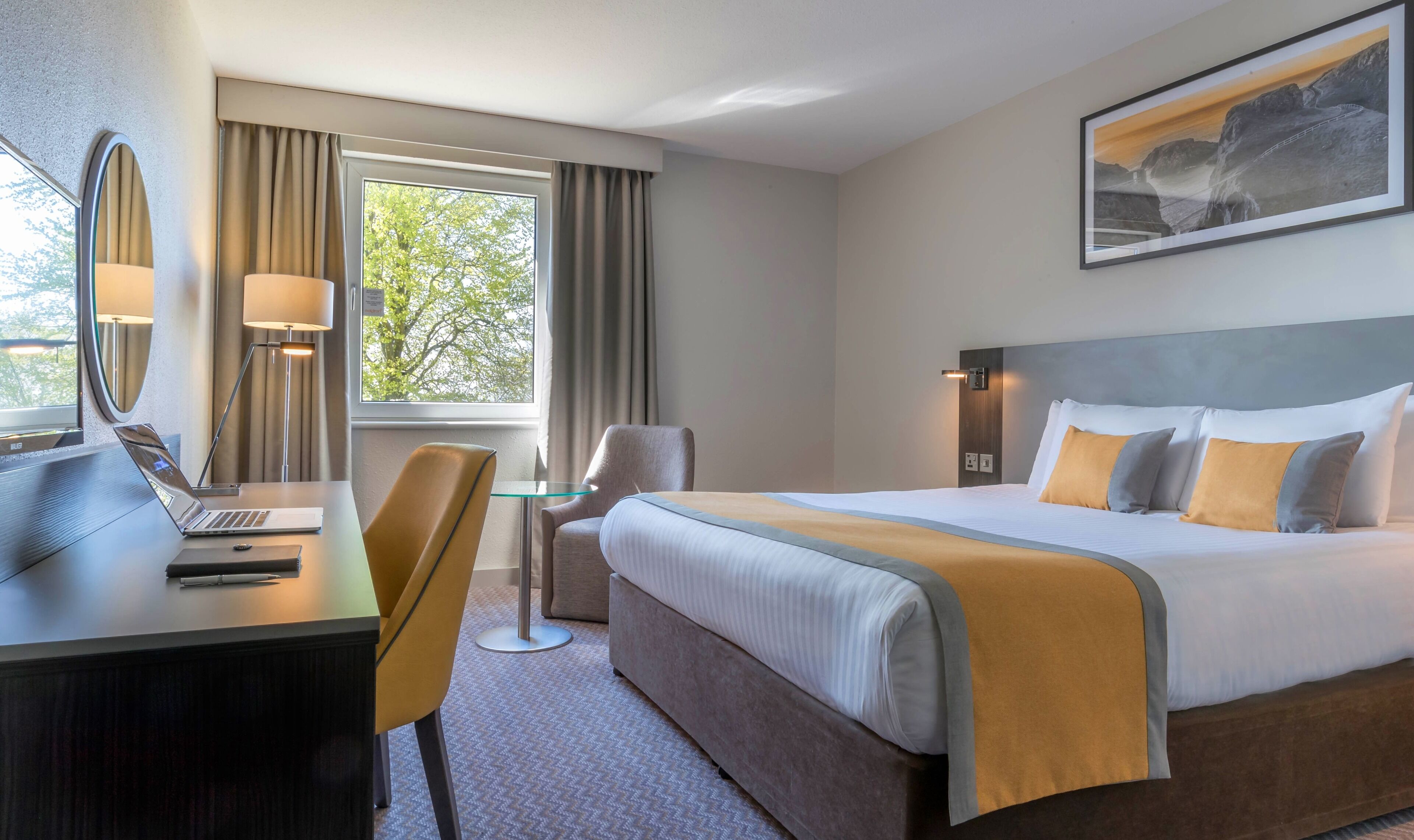 A hotel room with a bed, desk, chair, lamp, and a painting on the wall.