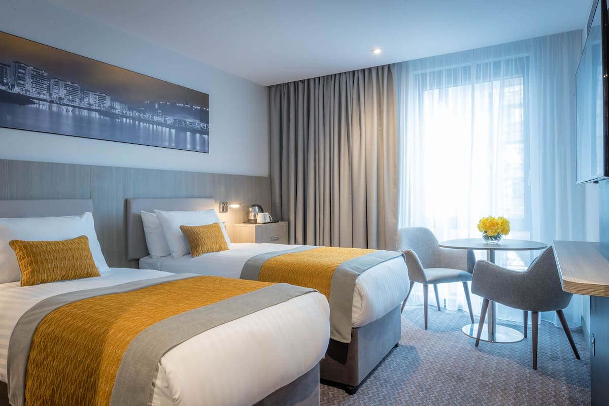 Ein Hotelzimmer mit zwei Betten, einem Schreibtisch, einem Stuhl, einem Tisch mit Blumen und Blick auf die Stadt.