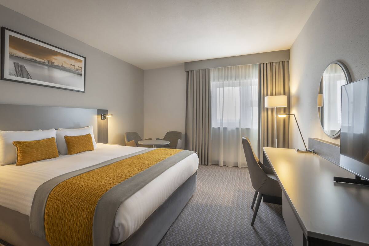 Ein Hotelzimmer mit einem großen Bett, einem Schreibtisch, einem Stuhl, einem runden Tisch, einem Fernseher und einem Bild an der Wand.
