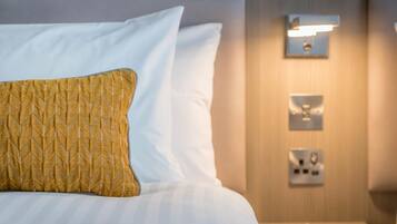 A neatly made bed with white and yellow pillows, a bedside table with a lamp, and a wall-mounted light fixture.
