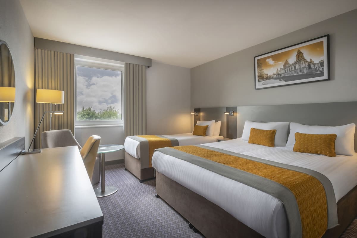 Ein Hotelzimmer mit zwei Betten, einem Schreibtisch, einem Stuhl, einem kleinen Tisch und einem Fenster mit Vorhängen.