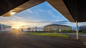 Un edificio moderno con estructura circular, un cartel de Belfast International Airport y un área verde al frente.